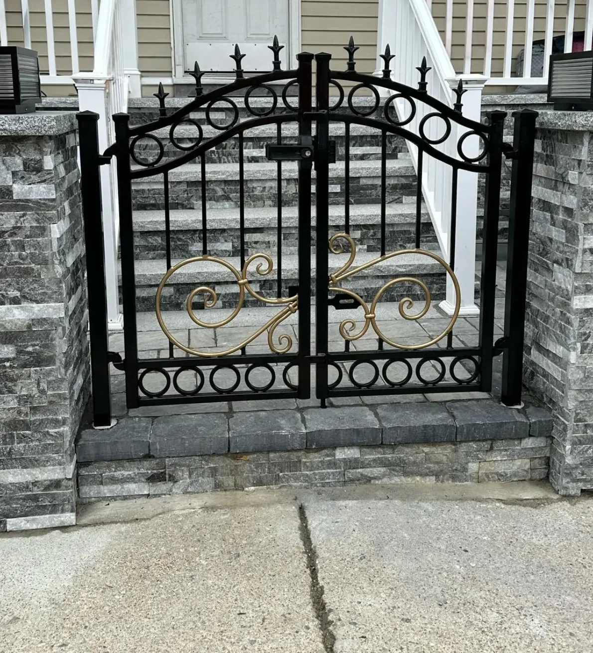 Custom double walk gate in black steel with gold scrollwork centerpiece, set between stone pillars